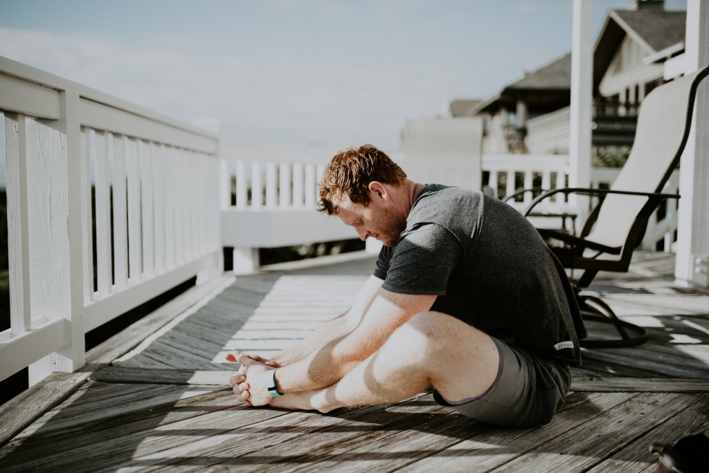 Man on a porch doing a butterfly yoga stretch.