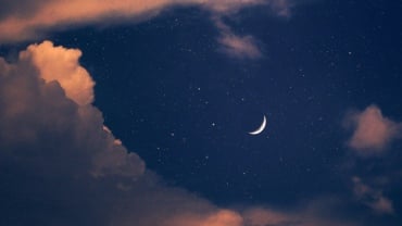 A close-up of a crescent moon in a navy-blue sky with pink-tinged clouds.