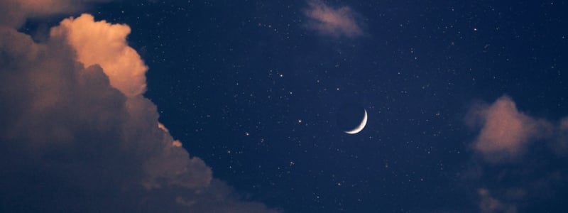 A close-up of a crescent moon in a navy-blue sky with pink-tinged clouds.