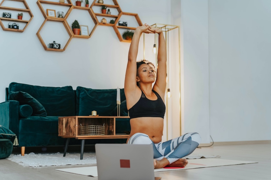 Woman in a black sports bra and grey leggings stretching on a yoga mat in front of an open laptop.