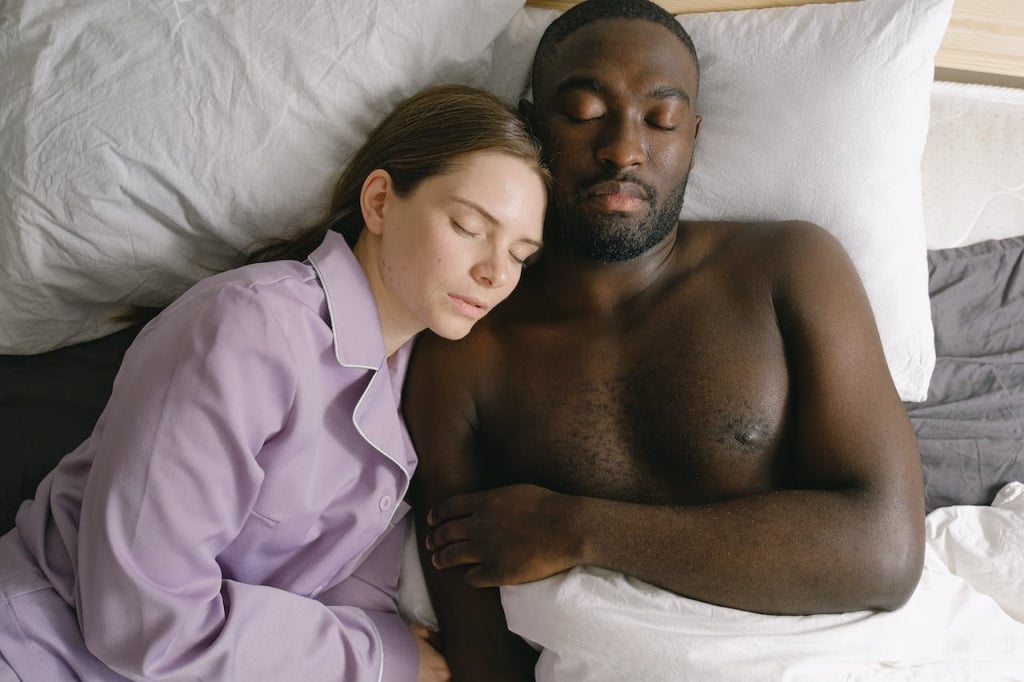 A man and a woman lying on their backs with their eyes closed in a bed with white sheets