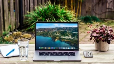 A laptop on an outdoor wooden table next to a glass of water; plants and shrubs are in the background