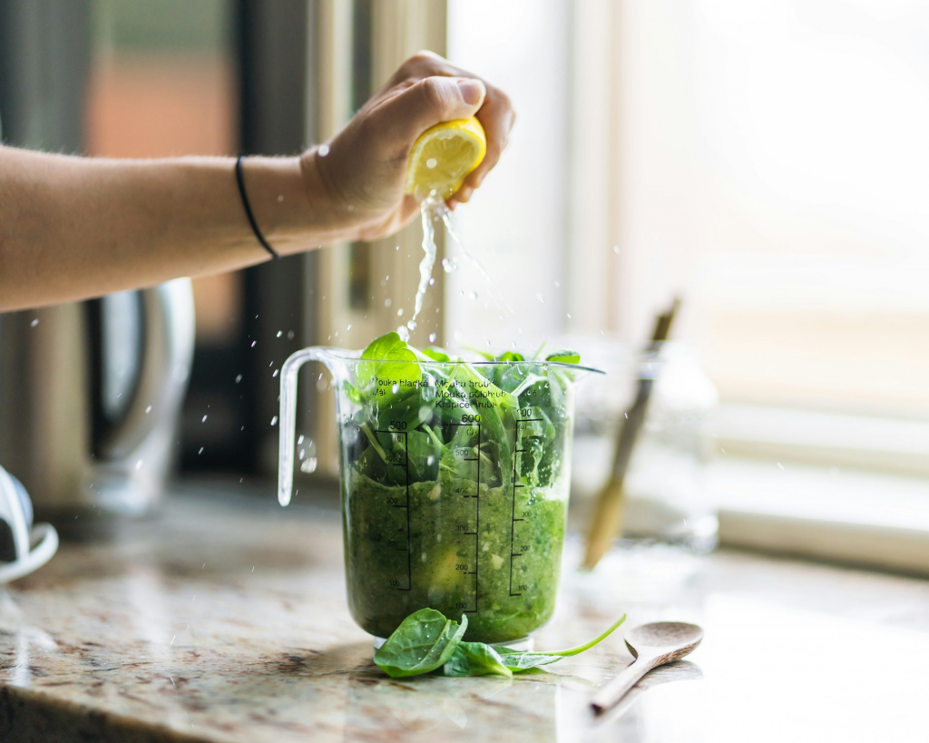 Ingredients for a green smoothie in a blender, with a hand squeezing a lemon into it.