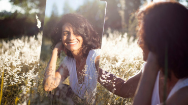 Woman sitting in a meadow and smiling at her reflection in a mirror she is holding