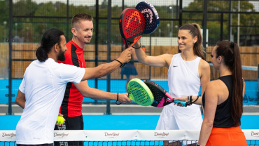 Four men and women on an outdoor Padel court holding up their padels.