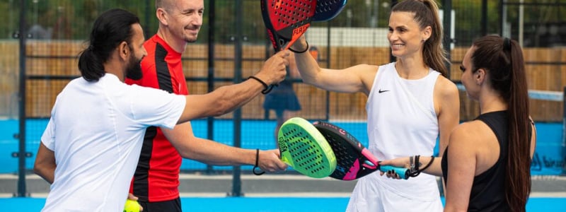 Four men and women on an outdoor Padel court holding up their padels.