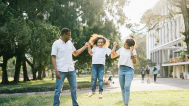 A man and a woman swinging a little girl by the arms in a sunny park.