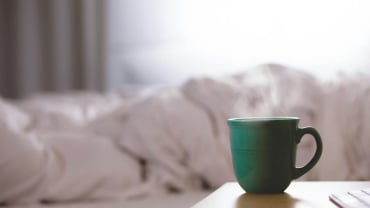 A steaming mug in the foreground with what looks like rumpled white bedsheets in the background.