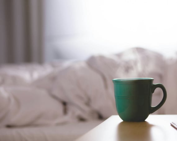 A steaming mug in the foreground with what looks like rumpled white bedsheets in the background.