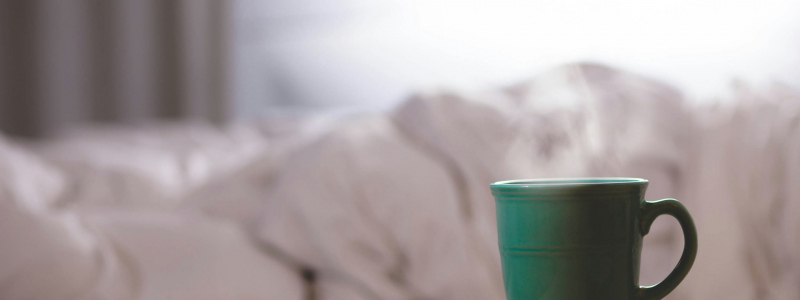 A steaming mug in the foreground with what looks like rumpled white bedsheets in the background.