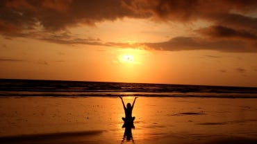 A silhouette of a person sitting on a beach with arms raised up against a sunset.