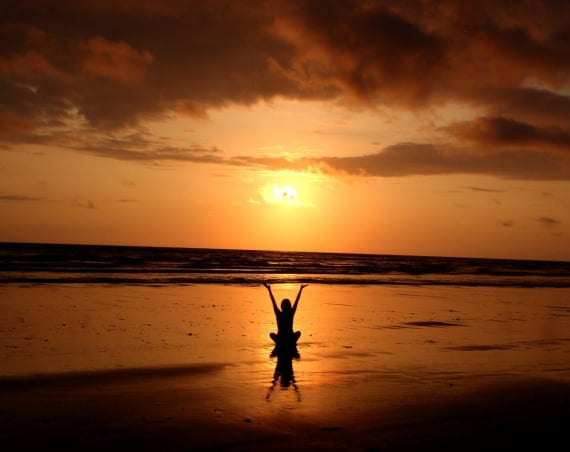 A silhouette of a person sitting on a beach with arms raised up against a sunset.