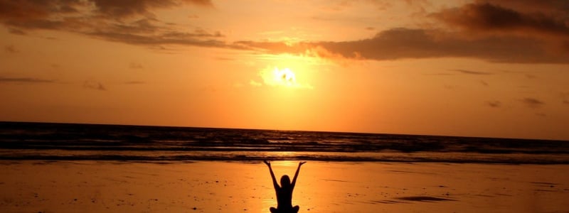 A silhouette of a person sitting on a beach with arms raised up against a sunset.