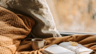 A notebook resting on a blanket with a leaf on top of it, in a brown shade that suggests autumn.