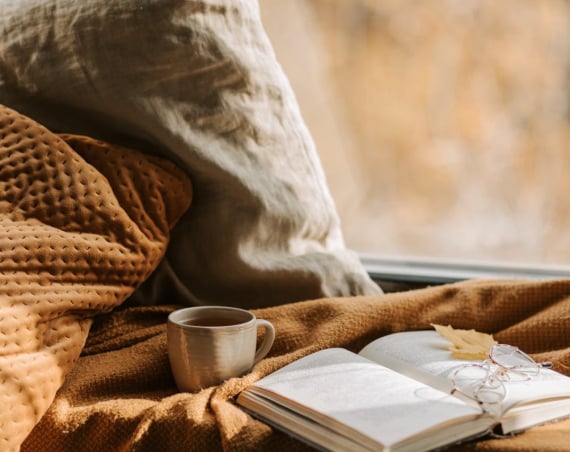 A notebook resting on a blanket with a leaf on top of it, in a brown shade that suggests autumn.