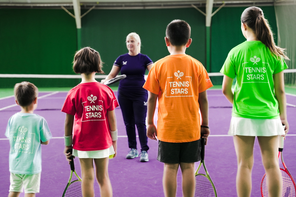 alt="Lezione di tennis per bambini su campo indoor con allenatore e giovani partecipanti Tennis Stars"